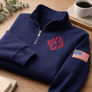 May include: Navy blue quarter-zip sweatshirt with a red embroidered logo and an American flag patch on the sleeve. The sweatshirt has a ribbed collar and cuffs, and a silver zipper. A cup of coffee sits in the background.