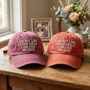 May include: Two baseball caps, one burgundy and one orange, with the embroidered text "I AM NOT LIKE OTHER GIRLS. I AM LIKE MY DAD." The caps are on a wooden table, with a framed photo and a vase of flowers in the background.