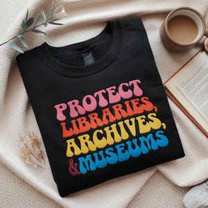 Könnte beinhalten: Schwarzer Pullover mit dem Text "PROTECT LIBRARIES, ARCHIVES, & MUSEUMS" in Regenbogenfarben.