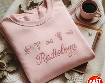 Puede incluir: Una sudadera de cuello redondo rosa claro con iconos bordados de líneas que representan una lámpara de escritorio, un martillo, un lazo y una caja torácica, con la palabra Radiology escrita en cursiva bajo los iconos, en hilo rosa a juego.
