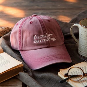 May include: A dusty rose baseball cap with the embroidered text "I'd rather be reading." The cap is sitting on a dark gray surface next to a book, a mug, and a pair of glasses.