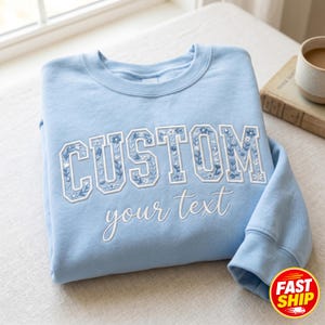 May include: A pale blue sweatshirt featuring the word "CUSTOM" in white, floral-patterned letters. Below, the words "your text" are written in a cursive font. The sweatshirt is folded on a white surface.