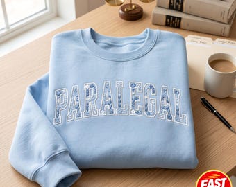 May include: A light blue sweatshirt with the word "PARALEGAL" in white, floral-patterned lettering. The sweatshirt is folded on a wooden surface, with law books, a coffee mug, and a scale of justice in the background.