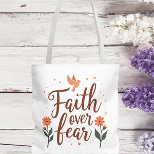 May include: White tote bag with the words "Faith over fear" in brown script, a flying bird, and orange flowers. The bag has a white handle and is set against a white wooden background with purple and white flowers.