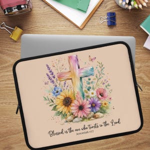 May include: A rectangular laptop sleeve with a light peach background featuring a watercolor illustration of a colorful cross surrounded by sunflowers, daisies, and lavender. The text reads: Blessed is the one who tursts in the Lord Jeremiah 17:7.