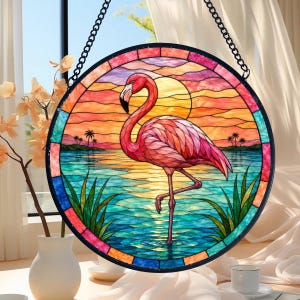 Puede incluir: Un atrapasoles circular de vidrieras que muestra un flamenco rosa de pie en agua azul. El fondo representa una puesta de sol con nubes naranjas y amarillas, un gran sol y siluetas de palmeras. La pieza tiene un marco negro y cuelga de una cadena.