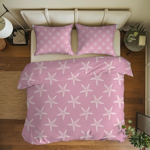Pode incluir: Um conjunto de roupa de cama rosa com um padrão de estrelas do mar brancas, incluindo uma capa de edredão e duas fronhas. As estrelas do mar são brancas e espaçadas uniformemente sobre o fundo rosa. A roupa de cama está numa cama branca.