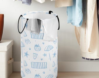 May include: A cylindrical white laundry hamper featuring a repeating blue line-art pattern of beach-themed icons including flip-flops, beach towels, umbrellas, starfish, coconuts, and sunscreen, with two black fabric handles at the top rim.