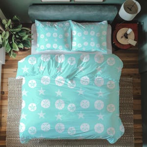 Op de afbeelding: Een turquoise dekbedovertrek en twee kussenslopen met een wit zeester- en zanddollarpatroon. De beddengoedset ligt op een bed met een groen hoofdbord en een houten vloer. Het thema is kust.