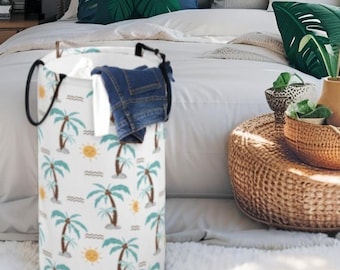 May include: A cylindrical white laundry hamper featuring a repeating pattern of teal palm trees and yellow suns. The hamper has two black fabric handles attached to the top rim and contains a pair of folded blue denim jeans inside.