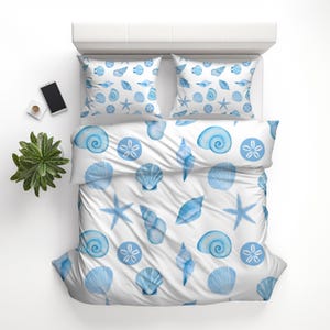 May include: White bedding set with a blue seashell pattern. The set includes a duvet cover and two pillowcases. The design features various seashells, starfish, and sand dollars in a watercolor style. A smartphone and a small plant are on the side.