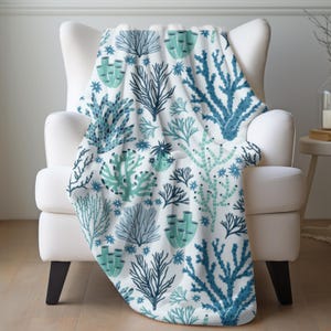May include: A white armchair draped with a throw blanket featuring a repeating pattern of stylized coral and seaweed in shades of blue and teal. The blanket's design includes various coral shapes and star-like accents, creating a marine theme.