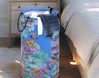 May include: A cylindrical laundry hamper featuring a vibrant underwater coral reef print with orange clownfish and blue tang fish. The hamper has a blue rim, black fabric handles, and contains white fabric and a pair of blue denim jeans.