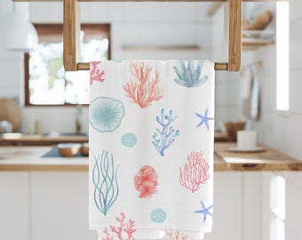 Puede incluir: Toalla de cocina blanca con un diseño de acuarela de coral, estrellas de mar y plantas marinas en tonos azules, rosas y verdes. La toalla está colgada en un toallero de madera.