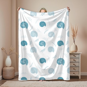 Op de afbeelding: Witte deken met een herhaald patroon van blauwe schelpen en takjes. De schelpen variëren in blauwtinten en hebben gedetailleerde spiraalontwerpen. De deken wordt omhoog gehouden, waardoor het volledige patroon en de zachte textuur te zien zijn.