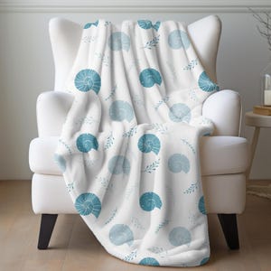 Op de afbeelding: Een witte deken met een patroon van blauwe schelpen en takjes, gedrapeerd over een witte fauteuil. De schelpen hebben verschillende tinten blauw. De deken ziet er zacht en gezellig uit. De fauteuil heeft zwarte poten.