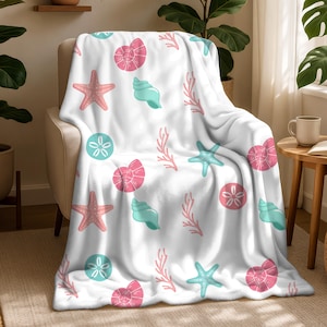 Op de afbeelding: Witte plaid met een herhalend patroon van roze zeesterren, schelpen en koraal, samen met turquoise schelpen en zanddollars. De plaid is over een beige fauteuil gedrapeerd, wat een kustthema creëert.