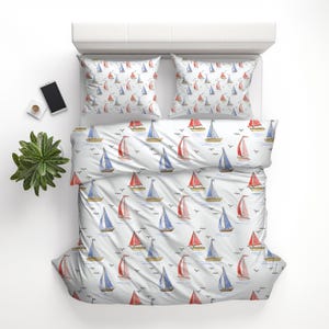 Puede incluir: Juego de cama blanco con un estampado de acuarela de veleros rojos, azules y amarillos. El juego incluye una funda nórdica y dos fundas de almohada. Un smartphone, una planta pequeña y una taza están al lado.