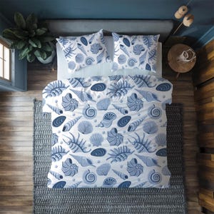 Puede incluir: Un juego de cama blanco y azul con estampado de conchas marinas, que incluye una funda nórdica y dos fundas de almohada, se muestra en una cama. El diseño presenta varias ilustraciones de conchas marinas en tonos azules. La cama está sobre una alfombra gris.
