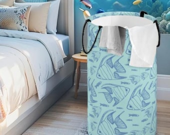 May include: A cylindrical laundry hamper featuring a light blue background with a repeating dark blue line art pattern of angelfish and smaller fish. The hamper has two black fabric handles and contains a white cloth draped over the top rim.