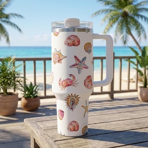 Peut inclure: Mug de voyage blanc avec une poignée, décoré d'illustrations à l'aquarelle de coquillages, d'étoiles de mer et d'un crabe. Le mug a un couvercle transparent et est posé sur une plage avec des palmiers.