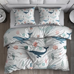 May include: White bedding set with a whale and sea life pattern. The duvet cover and two pillowcases feature blue whales, coral, and seaweed designs. The bed is made up with a white sheet and two small side tables.