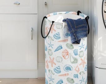 May include: A cylindrical laundry hamper featuring a white base with a repeating pattern of orange starfish and various blue and green seashells. The hamper has two black fabric handles and contains a pair of folded blue denim jeans inside.