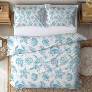 Pode incluir: Um edredom branco e duas fronhas com um padrão repetido de criaturas marinhas azuis claras, incluindo estrelas do mar, caranguejos, tartarugas e conchas. A roupa de cama está em uma cama branca.