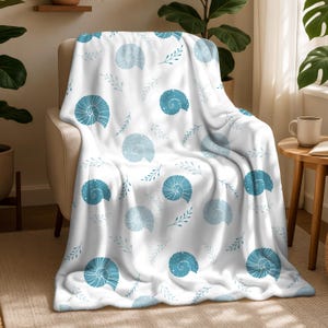 Op de afbeelding: Een witte deken met een herhaald patroon van blauwe schelpen en takjes. De deken is over een beige fauteuil gedrapeerd. De schelpen variëren in blauwtinten en hebben gedetailleerde spiraalontwerpen. De takjes zijn lichtblauw-grijs van kleur.