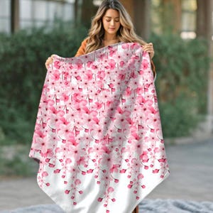 以下が含まれることがあります： ピンクの桜の花柄が繰り返される白いブランケット。花は様々なサイズとピンクの色合いで、下部に白い縁取りがあります。ブランケットは、花の模様を示すために提示されています。