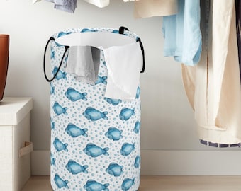 May include: A cylindrical laundry hamper featuring a white background with a repeating pattern of blue watercolour-style fish and small dots. The hamper has two black fabric handles and contains a few pieces of white and grey clothing inside.