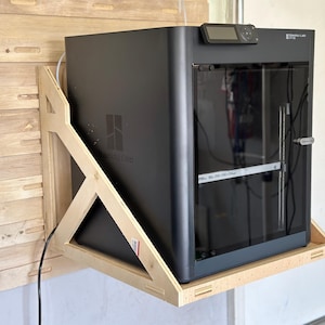 May include: A black 3D printer mounted on a wooden shelf. The printer has a glass door and a digital display. The shelf is attached to a wall with a wooden frame.