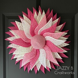 May include: A circular wreath featuring layered, pointed petals made of mesh fabric in shades of hot pink, light pink, and cream. The center consists of a round, spiraled dome of pink rope. The text Jazzy B Craftworks is visible in the bottom right.