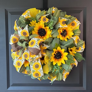 May include: A decorative wreath featuring artificial sunflowers with yellow petals and dark centers, interspersed with green mesh ribbon, yellow mesh accents, and a patterned fabric ribbon featuring a sunflower print and solid brown edges.