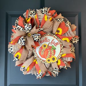 May include: A decorative wreath featuring orange, brown, and leopard print mesh ribbons, accented with yellow sunflowers. A central circular sign displays the text "hello fall" above a pumpkin illustration with leopard print and sunflower details.