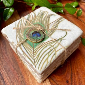 May include: Set of four square coasters with a peacock feather design. The coasters are made of stone and have a white background with a green and gold peacock feather design. The coasters are tied together with twine and have a small round decorative piece with a peacock eye design.