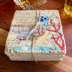 May include: Set of four coasters featuring a nautical theme with seashells, coral, and a blue and white patterned design. The coasters are made of natural stone and have a rustic, distressed look. The text "Double the Rule" is printed on the coasters.