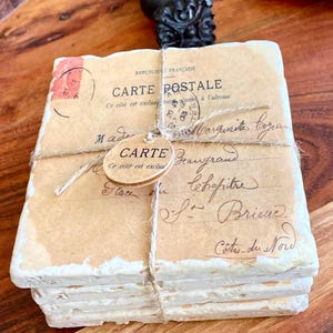 May include: A stack of four vintage postcards tied together with twine. The postcards are cream colored with white edges and have a faded look. The top postcard has French text that reads 