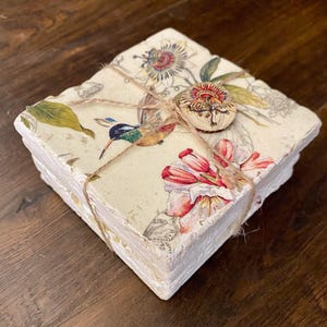 May include: Set of four square coasters with a floral and hummingbird design. The coasters are white with a distressed finish and are tied together with twine. A wooden button with a floral design is attached to the twine.