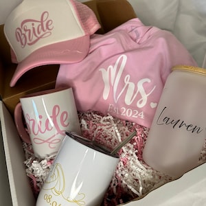 May include: A pink and white bridal gift box containing a pink trucker hat with the word "Bride", a pink t-shirt with the text "Mrs. Est 2024", a white mug with the word "Bride", a white tumbler with a gold design and the date "08.04.2024", and a clear glass with a pink lid that says "Lauren".