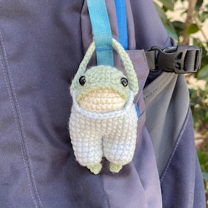 May include: A green and white crocheted frog plush toy hanging from a blue and gray backpack.