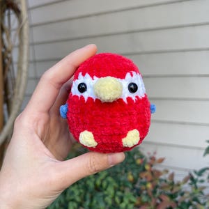 May include: A small, red crocheted bird with white eyes, a yellow beak, and blue wings.