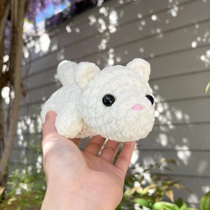 May include: A handmade, white crochet bear with black eyes and a pink nose. The plush toy is held in a hand, showcasing its soft, textured appearance. The bear has small, rounded ears and a simple, endearing design.