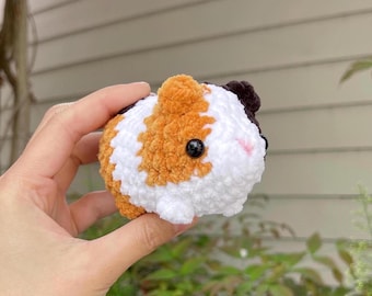 May include: A crocheted stuffed guinea pig toy with white, orange, and black fur. The toy has black eyes and a pink nose.