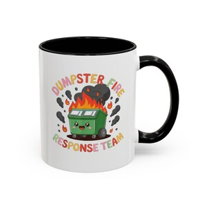 May include: A white ceramic mug with a black interior and handle. The mug features a cartoon illustration of a green dumpster on fire, with the text "DUMPSTER FIRE RESPONSE TEAM" in colorful letters.