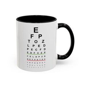 May include: A white ceramic mug with a black interior and handle. The mug features a classic eye chart design in black, with the letters arranged in descending sizes. The mug is a standard size for tea or coffee.
