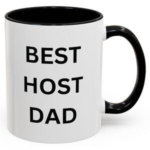 Pode incluir: Caneca de café de cerâmica branca com borda e alça pretas. A caneca tem o texto "BEST HOST DAD" impresso em preto.