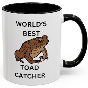 May include: White ceramic mug with a black rim and handle. The mug has the text "WORLD'S BEST TOAD CATCHER" printed on it. A cartoon image of a brown toad is in the center of the mug.