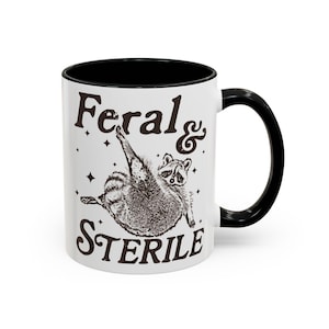 May include: White ceramic mug with a black interior and handle. The mug features a raccoon illustration with the words "Feral & Sterile" in brown text. Small star graphics are scattered around the raccoon.