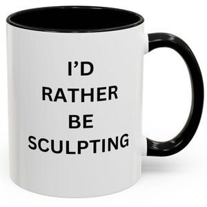 Op de afbeelding: Witte keramische mok met een zwarte rand en handvat. De mok heeft zwarte tekst met de tekst "I'd rather be sculpting".